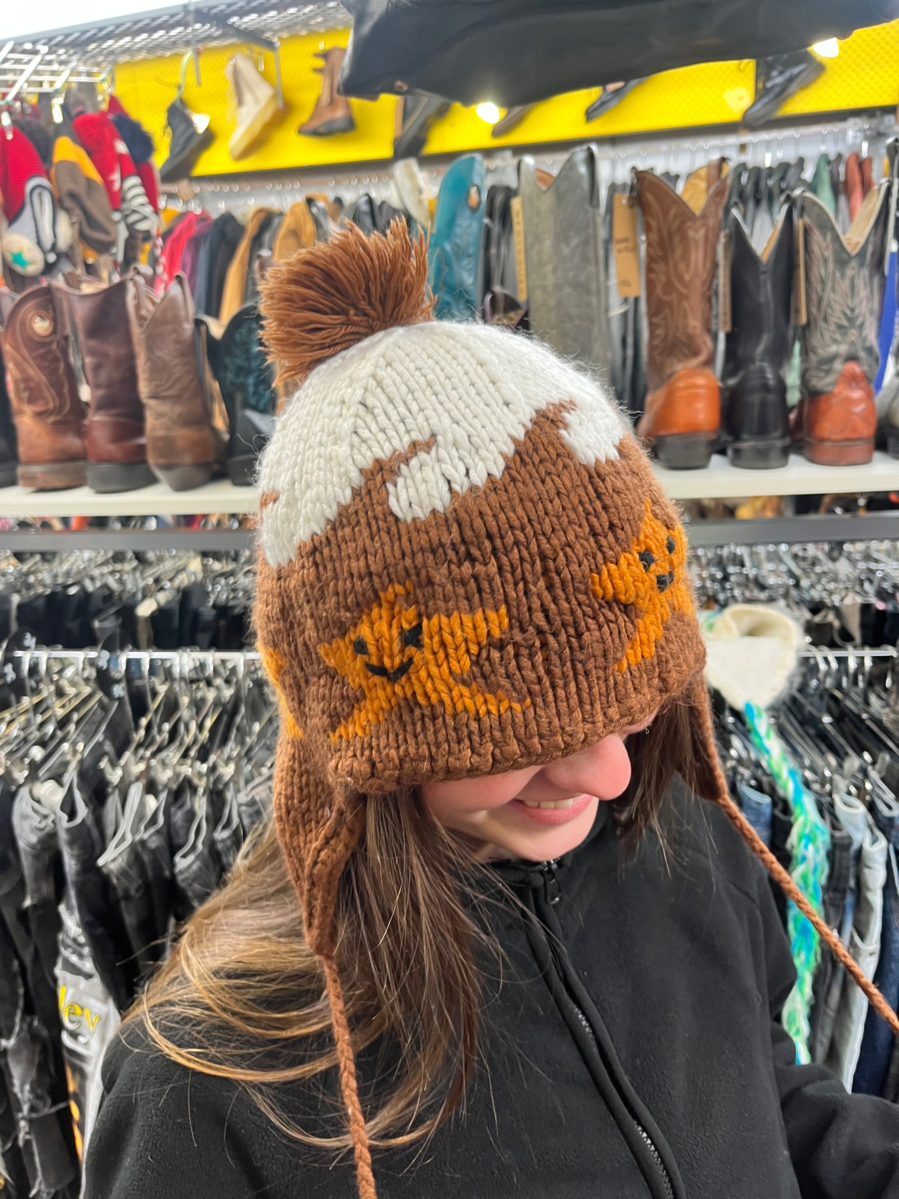 Side view of a person wearing a brown and white knit earflap beanie decorated with a snow-capped mountain pattern and smiling orange star graphics.