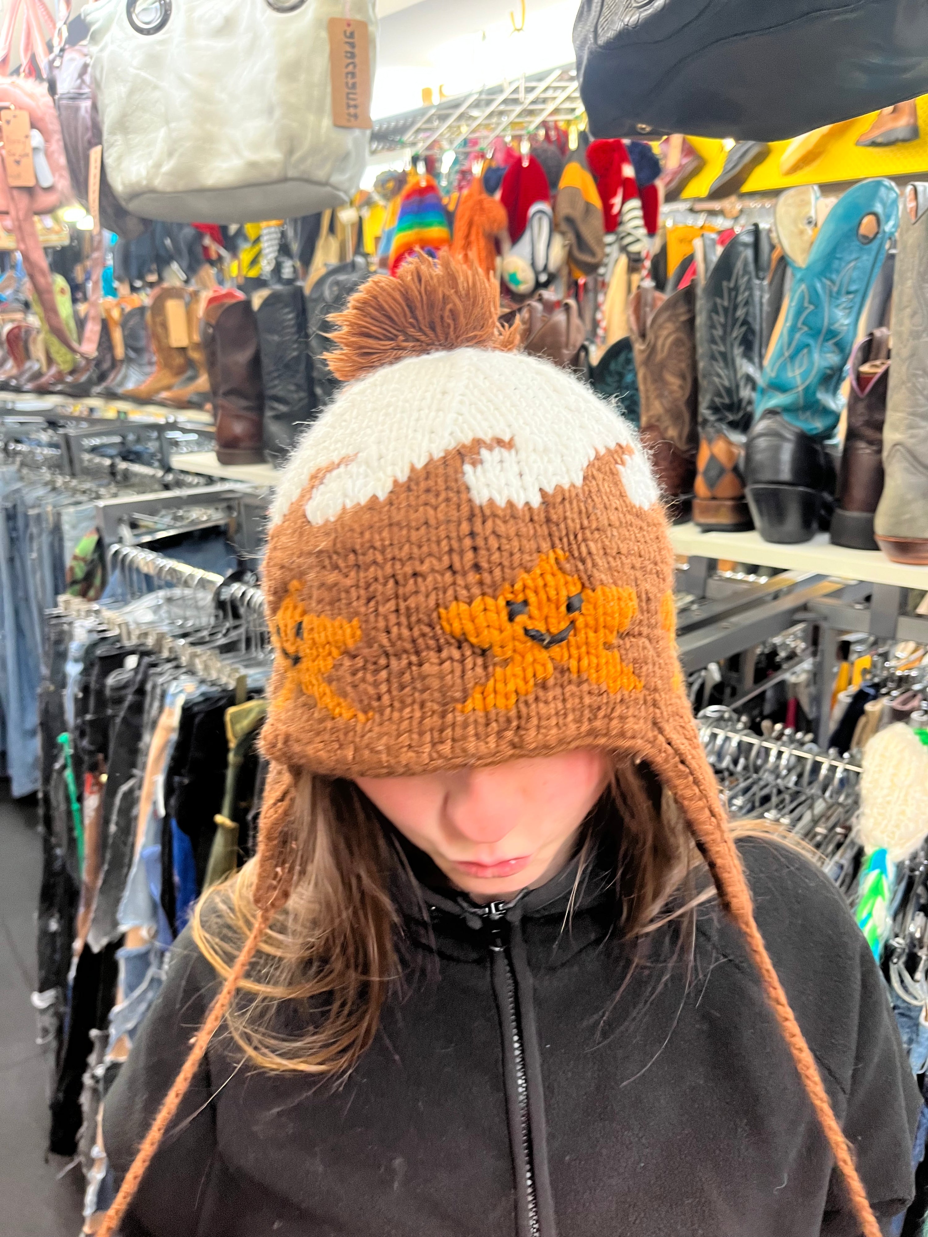 Front view of a vintage chocolate brown hand-knit winter hat featuring a fluffy top pom-pom, long side ties, and a pattern of happy, smiling cartoon stars.